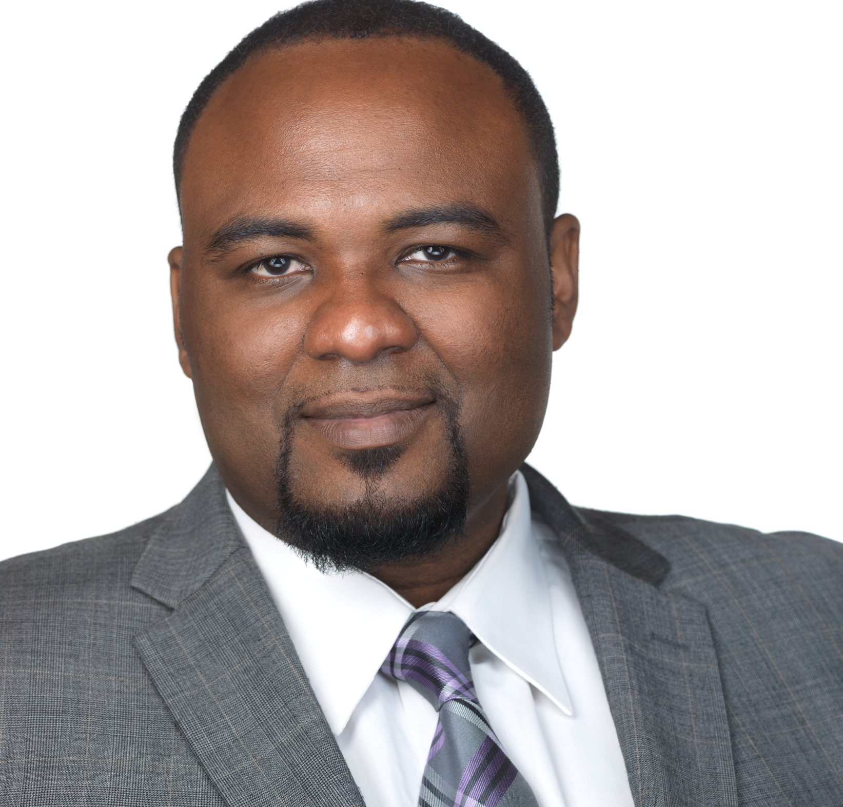Black man with short hair and goatee wears grey suit and smiles against white background.