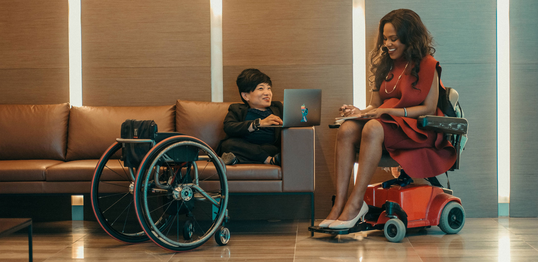 A woman using a wheel chair takes notes while speaking to a woman sitting on a couch using a laptop, with her wheelchair nearby.