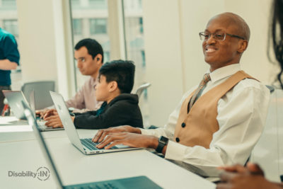 Man smiles while typing on computer. Next to him are two others working and one is a wheelchair user.
