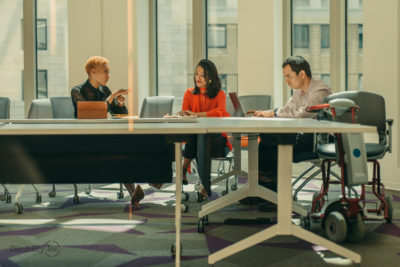 Three people sit in a board room. One woman signs ASL to another woman and the man is focused on his laptop.