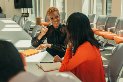 Two women sitting at a board table, one person is smiling and signing ASL.