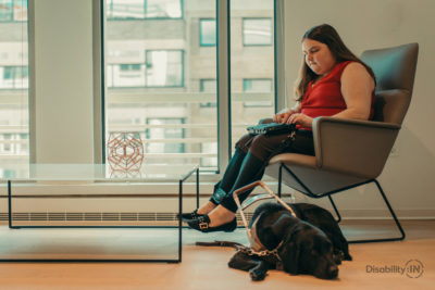 Woman sits on chair taking notes on braille notetaker. Guide dog sits on floor beside her.