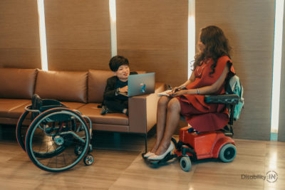 Two women meet informally in a corporate hallway. One woman is a wheelchair user and works on the couch, the other woman is on a motorized chair.