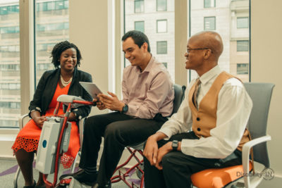 Three people looking at an iPad. Person in center uses a motorized scooter.