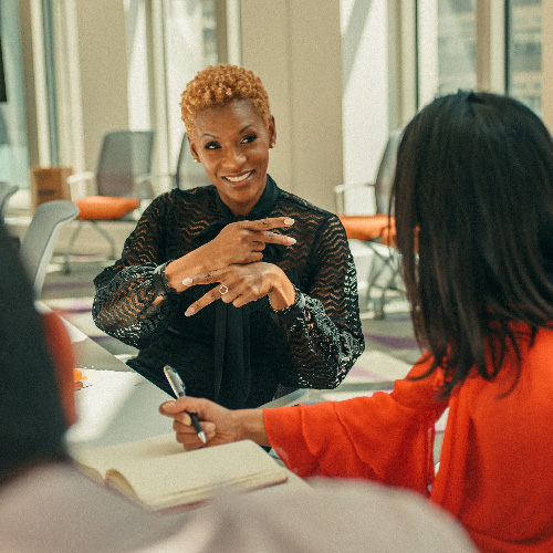 2021 Year in Review_In the News FastCo A black woman with short hair and black blouse signs to her colleague in red top while sitting at a conference table.