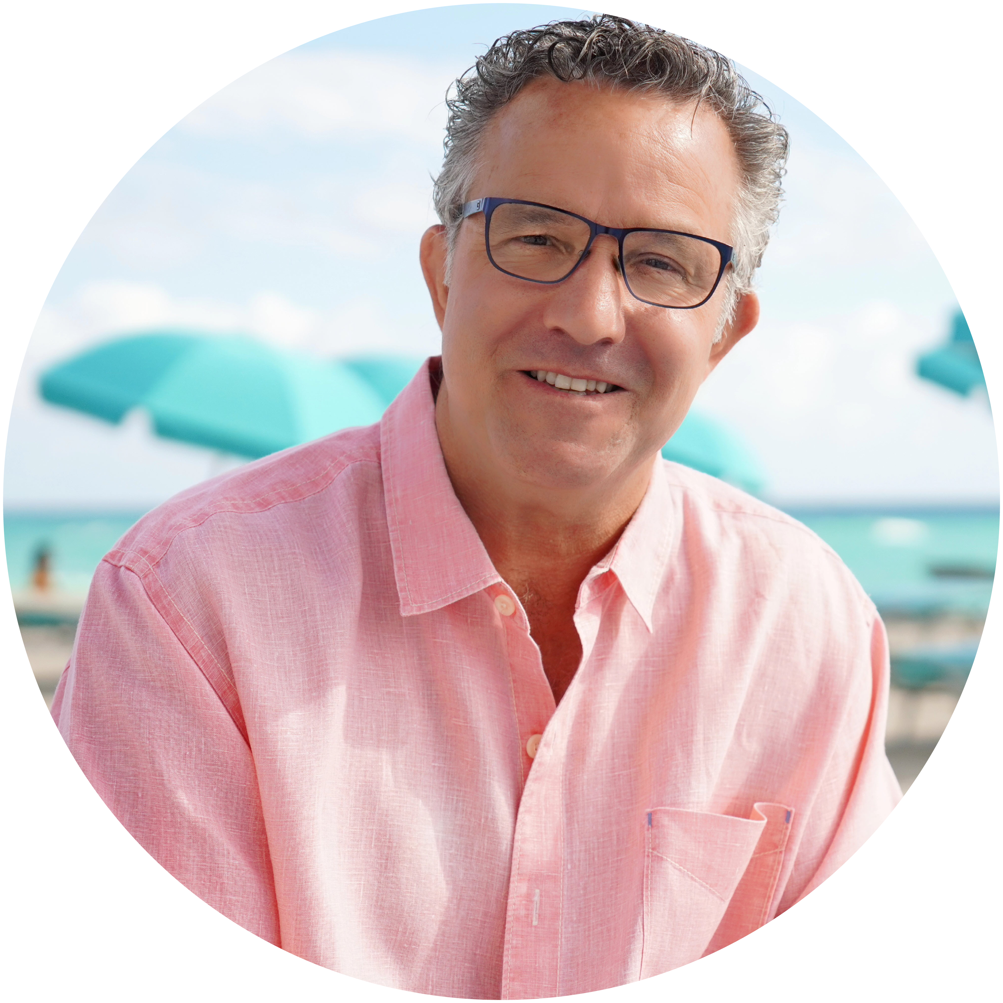 DOBE Semi-Finalists-Andrew Houghton Andrew Houghton, a white male with short grey hair and glasses wears a pink collared shirt against a beach background.